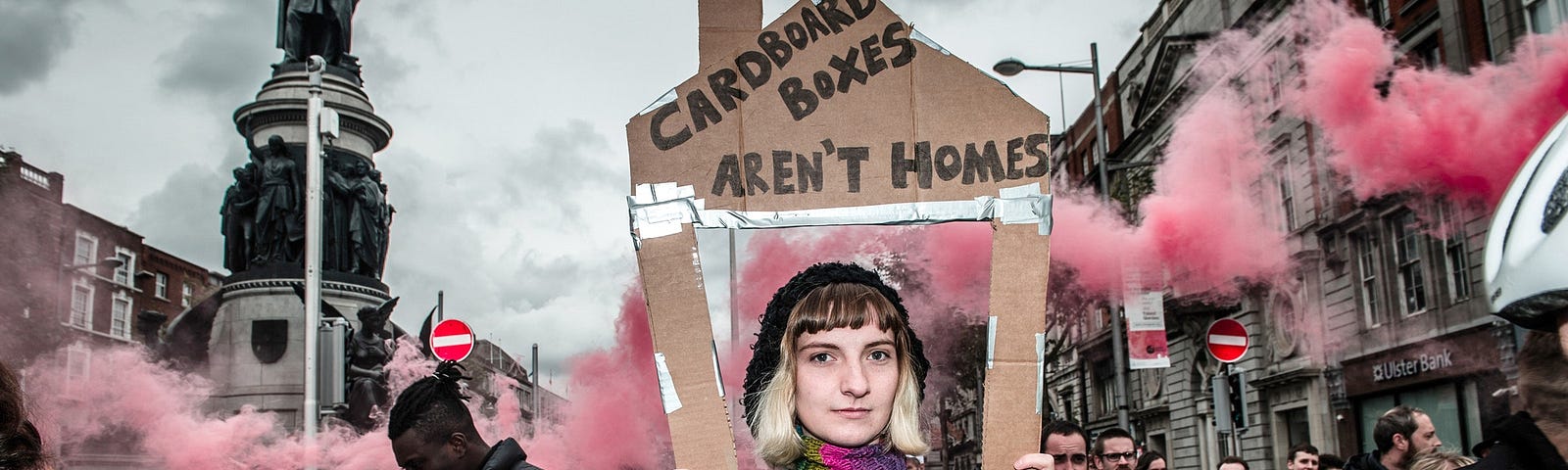 A woman holds a cardboard house with pink smoke behind her