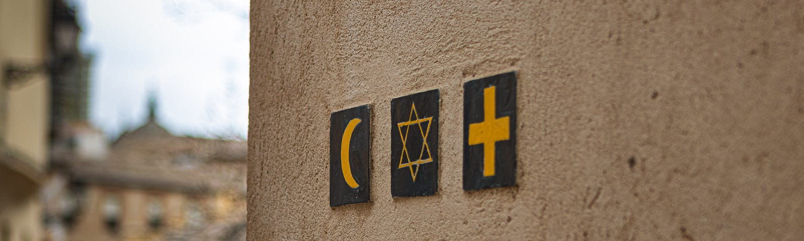 The crescent moon symbol of Islam, the star of David symbol of Judaism, and the cross symbol of Christianity on the side of a brown, mud wall.