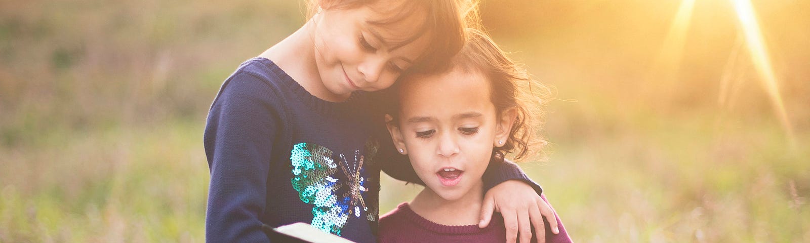 The image depicts two young children outdoors during what appears to be late afternoon, given the warm, golden sunlight in the background. The older child, wearing a navy-blue sweater with sequin embellishments and white leggings tucked into brown boots, is embracing a younger child from behind and holding an open book in front of them both. The younger child, in a maroon dress adorned with a white unicorn and matching maroon tights, is attentively looking at the book.