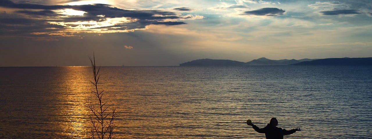 Person by sunset-lit lake as if in prayer, arms open wide.