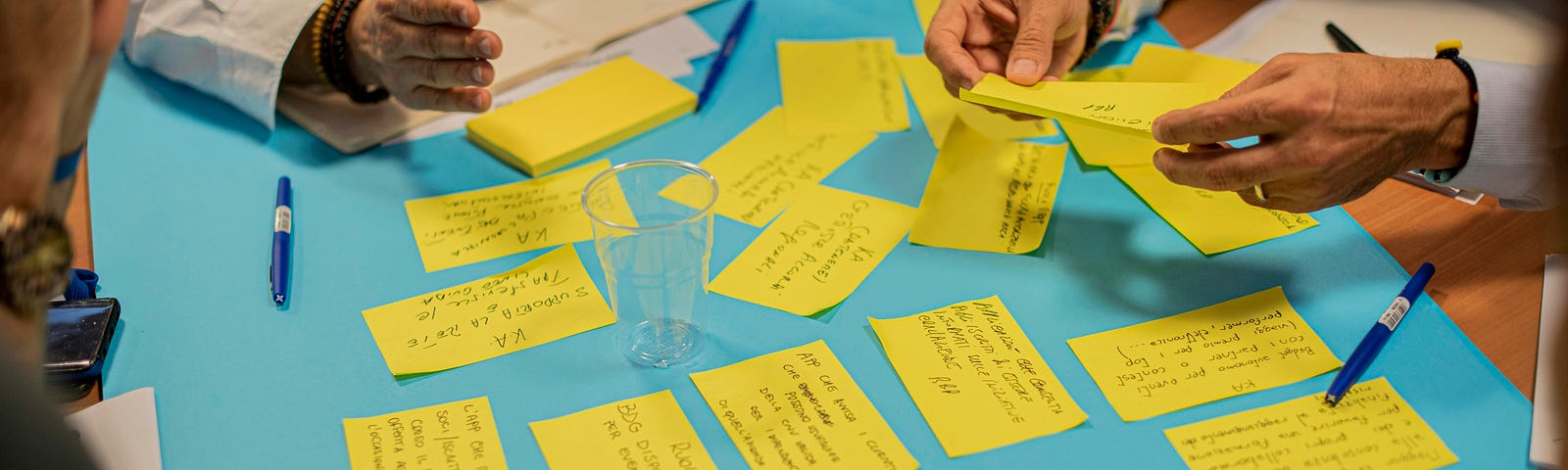 A group of people around a pale blue table are writing and exchanging yellow post it notes, a writers workshop.