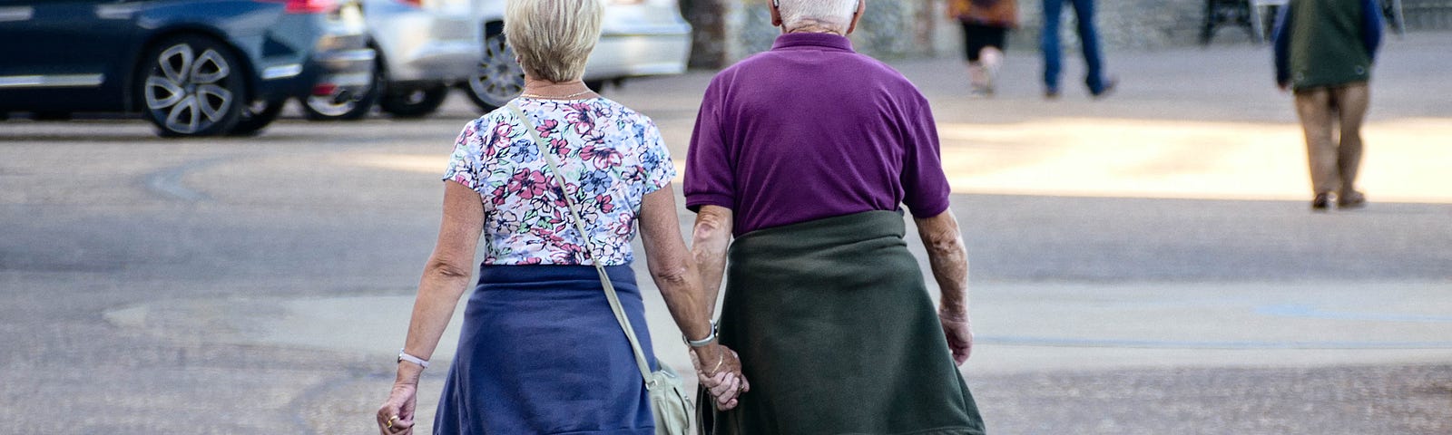 Two older people walking and holding hands in a city area.