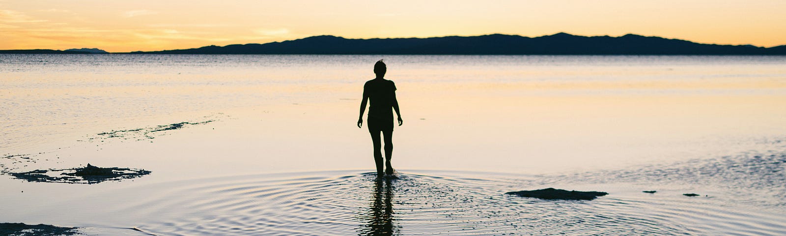 A woman wading out into the sea, with a mountain range in front of her, covered with a sunlit glow.