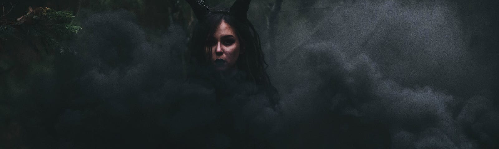 Overly dark image with greyish-black smoke and a face of a woman with devil hors behind the smoke.