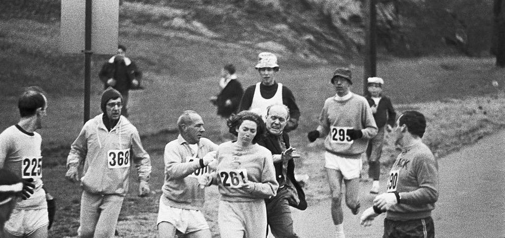 Black and white photo. Kathrine Switzer became the first woman to run the Boston Marathon. Here she is pictured making history while being attacked by the race organizer.