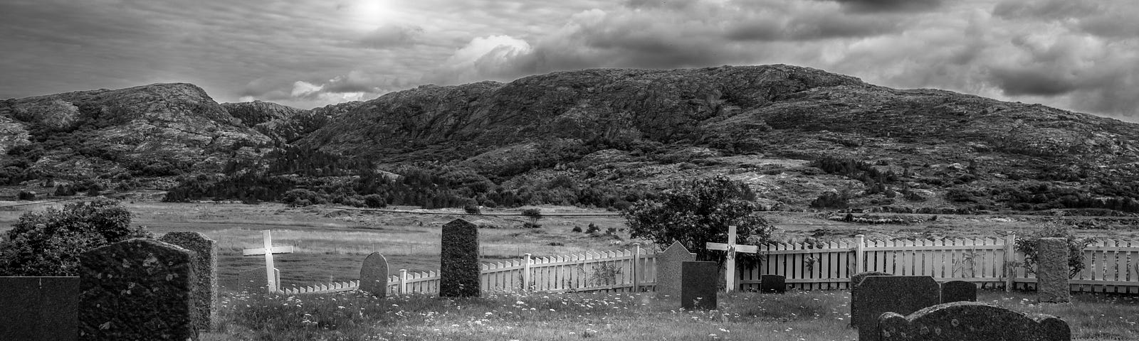graveyard with tombstones and crosses, a white fence and hills in the background