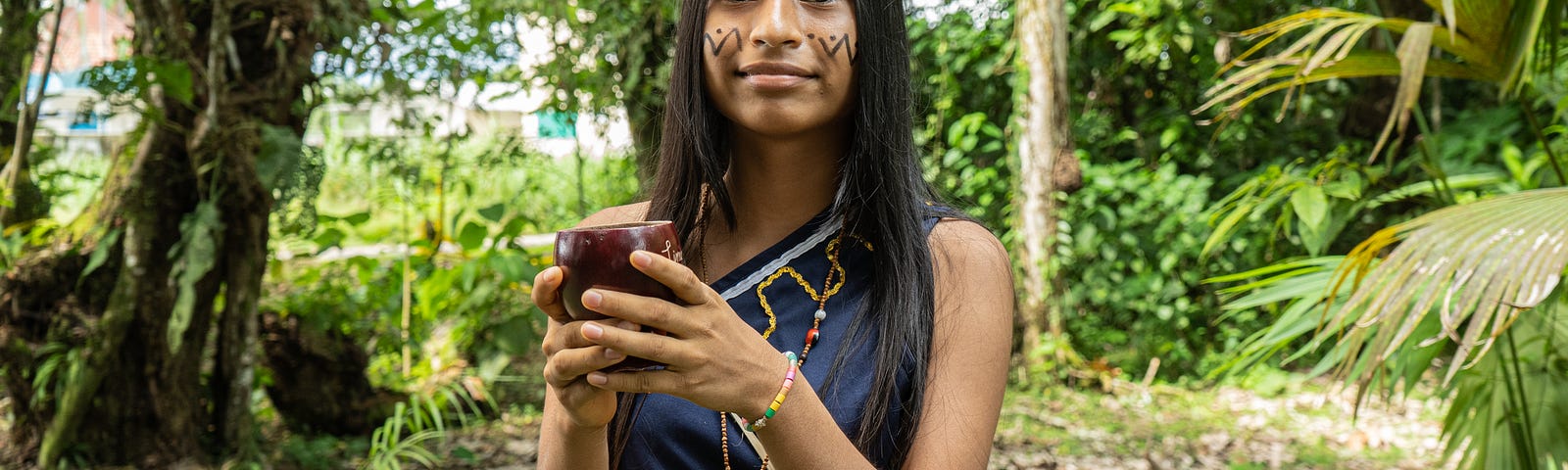 Amazon indigenous young woman in traditional clothing holding a cup and surrounded by nature.