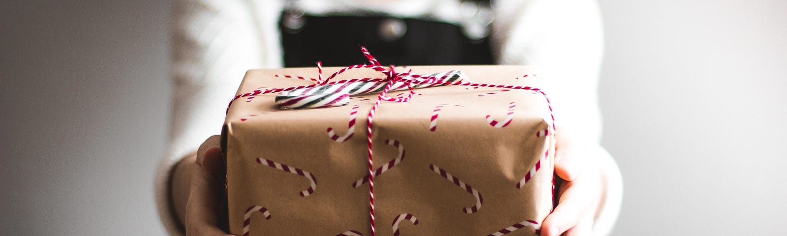 A woman dressed in a whiite shirt and black overalls hold out a small wrapped gift. The giftwrapping is brown with candy canes on it and a thin red and white ribbon wrapped around it.