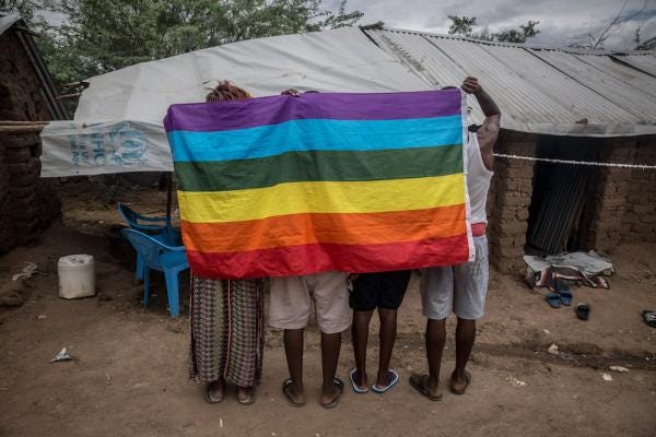 People stand behind a rainbow flag You can see the backs of their legs. A low tarp-covered building is in the background.