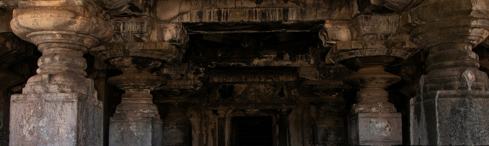 A passageway built of carved sandstone, its originally light colour stained grey or black in places with the passage of time, is flanked by massive decorated pillars, two on each side. At the end of the passage, a black door closes off a crypt beyond.