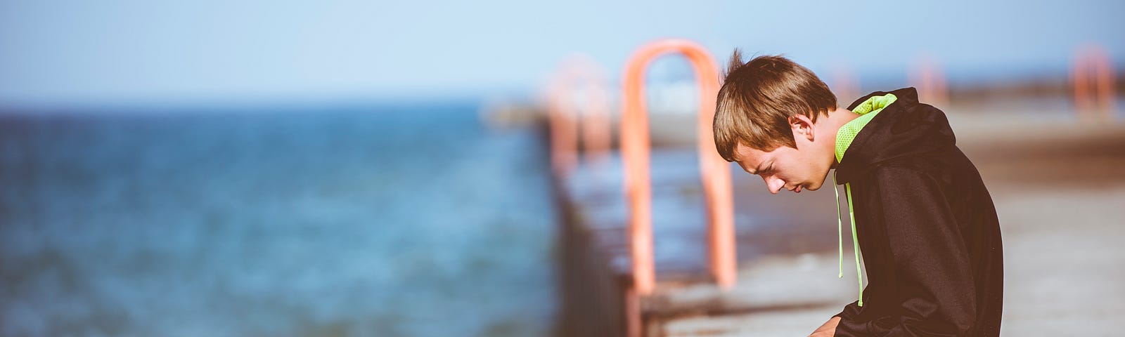 A very indoor-kid-looking indoor kid —a  boy, about thirteen or fourteen — sits on a dock reading instead of swimming.