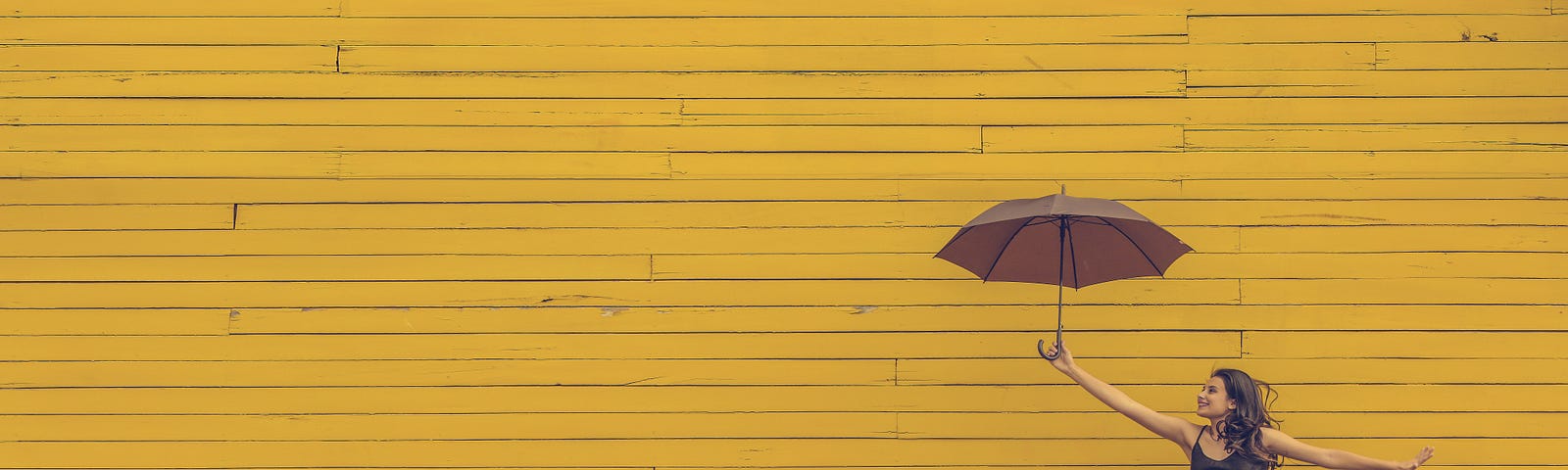 A woman jumping in front of a yellow wall while holding an umbrella.