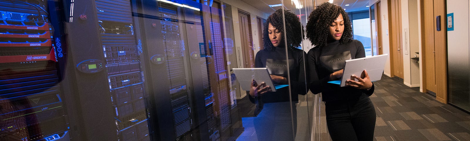 woman looking at a laptop in her hands standing next to a glass-walled room full of what appears to be computer racks
