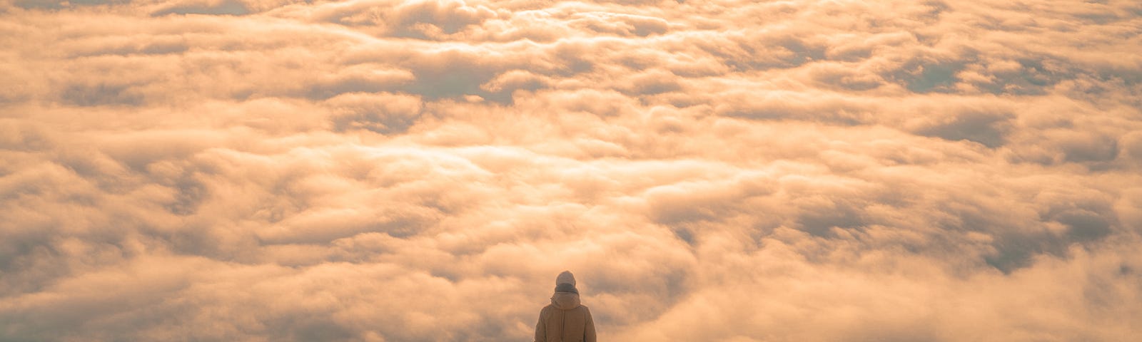 A person stands on the summit of a mountain, a ways off from the camera, looking out over a sea of clouds just below the mountain’s peak, with mountaintops rising through the clouds off in the distance.