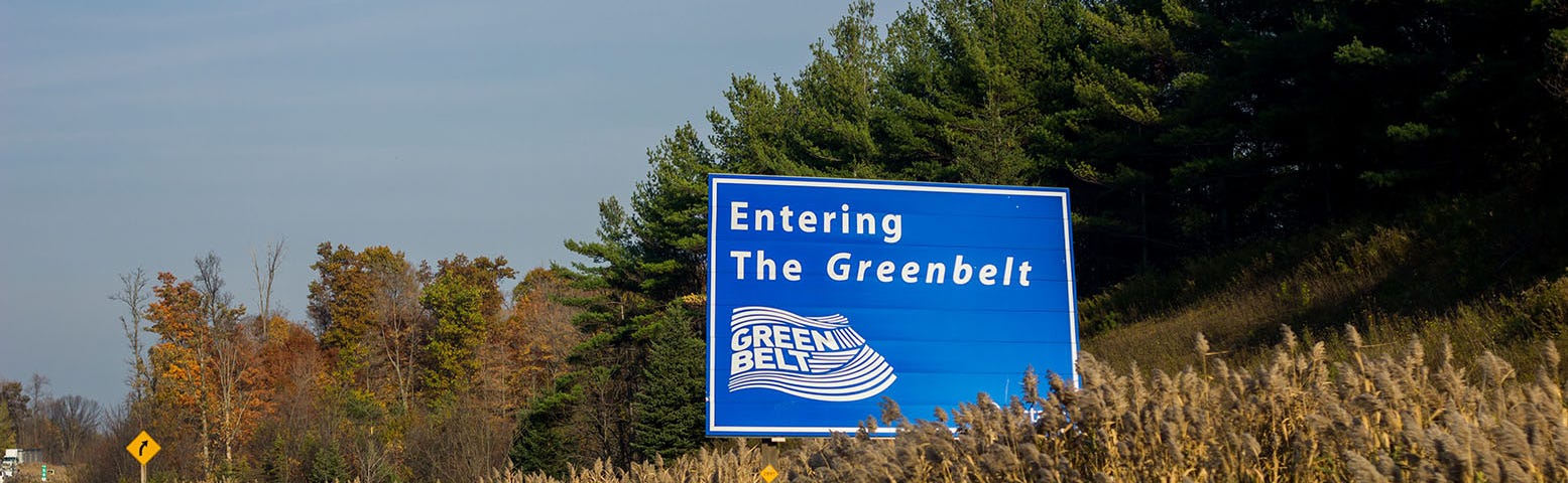 A road sign informs drivers they are entering the Greenbelt in Ontario. The highway is near a forest on a beautiful fall day.