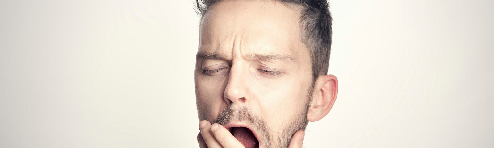 The image shows a man yawning with his eyes partially closed. He has short, styled hair and is wearing a white bathrobe. His hand is covering his mouth as he yawns, and the background is neutral and softly lit.