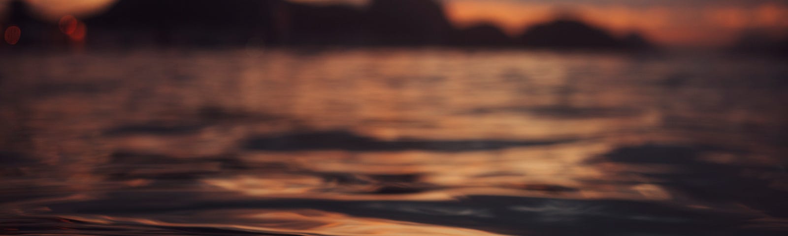 Closeup image of water, with silhouettes of mountains in the background. Image seems to have been taken in the setting sun.