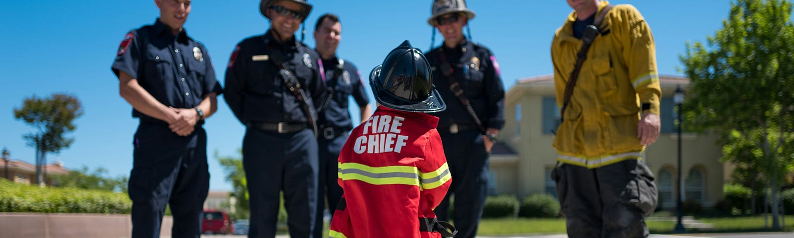 Young child in a Fire Chief costume with some firemen listening.
