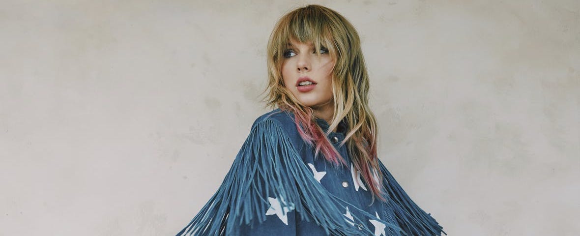 Taylor Swift looks coyly aside before a neutral background, wearing a fringey denim top with star patches