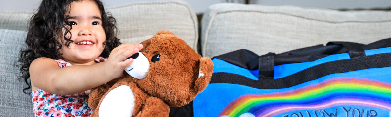 A smiling child sits on a couch holding a teddy bear. Next to her is a duffel bag decorated with the phrase, “Follow your dreams.”