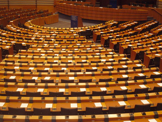 A close-up image of long, uninterrupted wooden desks and chairs that form a semi-circle. This is the European Parliament’s Hemicycle in Brussels.