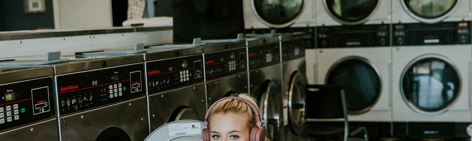 Cute young woman in a laundry mat with a clothes basket on her hip and headphones on her head.