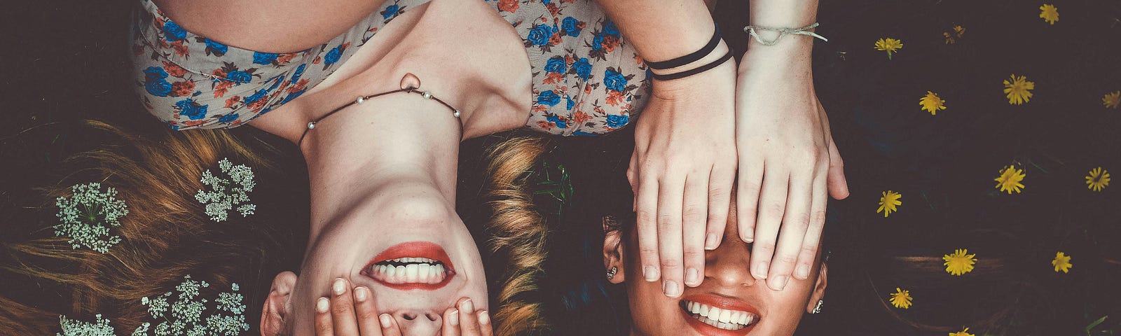 Two girls are lying next to each other. Their heads are next to each other but their bodies are in opposite directions. They are covering each other’s eyes with their hands and smiling.