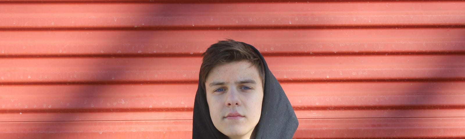 a youth in a dark hoodie standing against a reddish wall