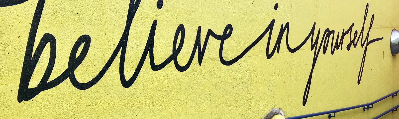 A kid in a yellow and black hoodie looks up at a yellow wall with big black words in script that says: “believe in yourself.”