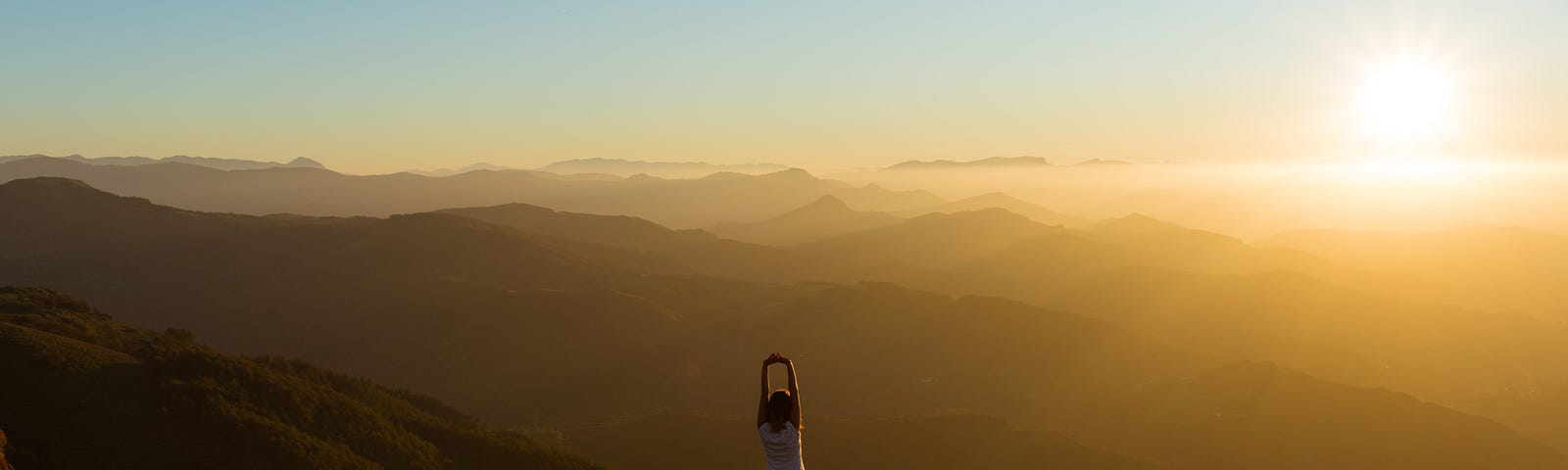 mulher de costas, em cima de uma montanha em postura de yoga olhando pro horizonte, que mostra mais montanhas e por do sol