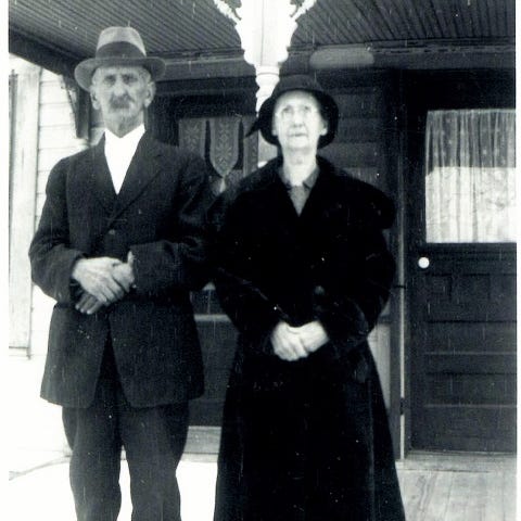 Two elderly people standing before a frame house with curtains