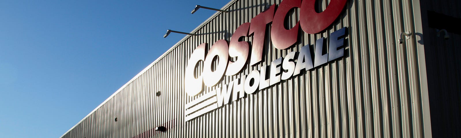 A massive white Costco warehouse looms across a bright blue sky, the name Costco Wholesale spelled out in large red and blue capital letters.