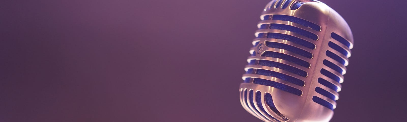 photo of an old-style microphone with silver metal outer and open lines in the structure of the device. The background is dark and blurry, with a clearly lit blurry spotlight shining where the mic is placed.
