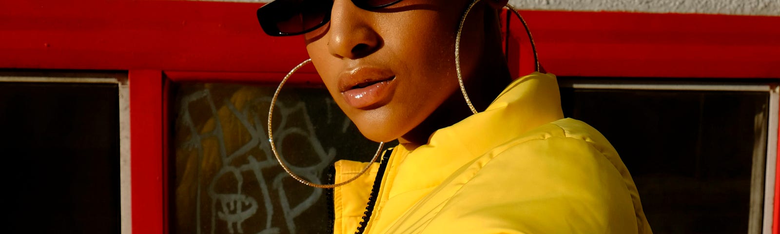 A lady with short black hair wears a yellow puffy jacket, extra large hoop earrings, and stylish dark glasses.