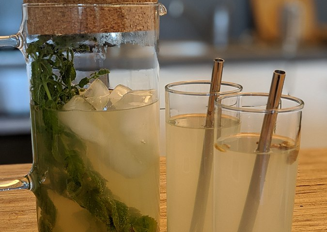 A photograph of a large glass pitcher with green herbs and lemonade with a light brown cork lid placed by two glasses filled with lemonade, and a metal straw in each glass. The glasses and pitcher are sitting on a surface made of very thin strips of wood joined closely together that are similar in color to the cork lid.