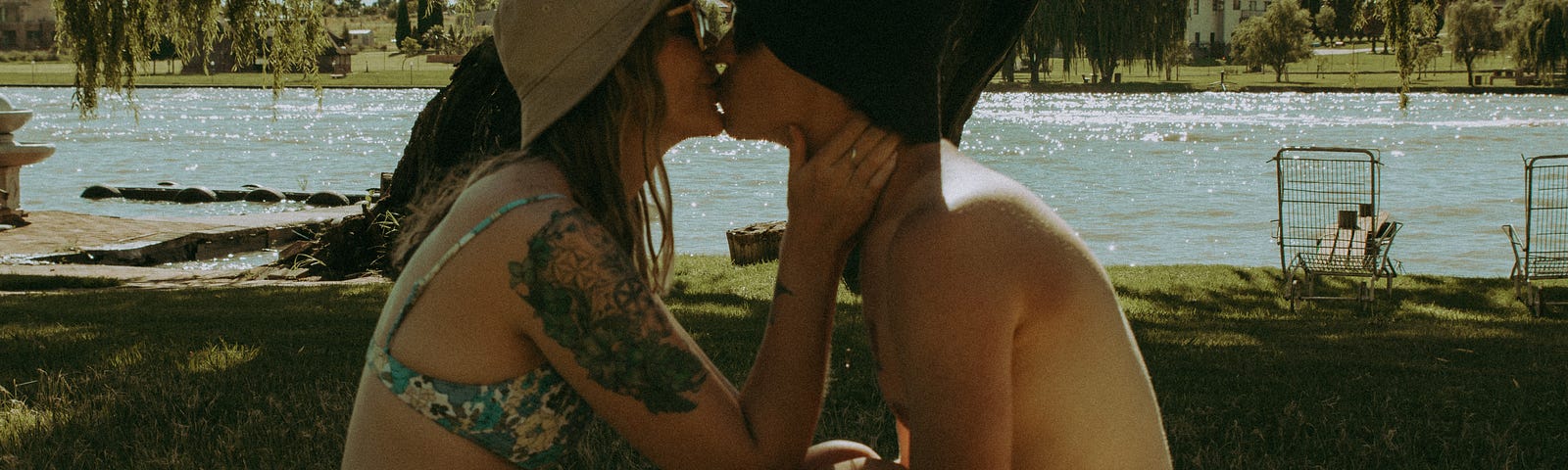 A man and woman sit on a blanket next to a river. They are facing each other with the woman’s legs over the man’s. They are kissing. Both are in swim suits and wearing hats. It looks hot and sunny.