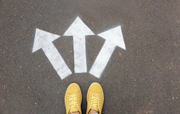 A pair of feet in yellow canvas sneakers standing in front of 3 arrows painted on the group that point in different directions.