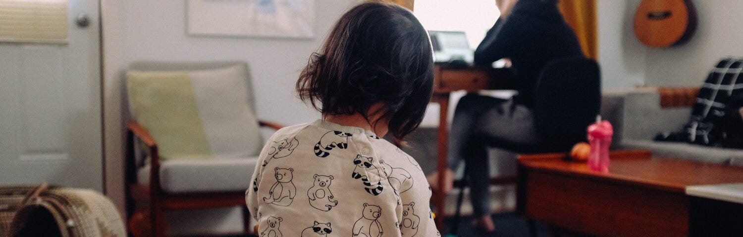 A child in the foreground, with parent working on laptop in background