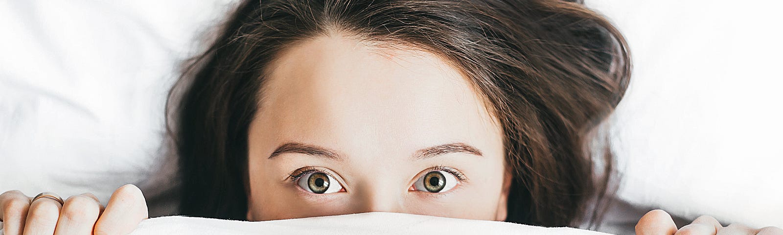 A brunette young woman pulls her bedsheet up to her nose level. We only see her two hands and her face from the mid-nose up. Insufficient sleep is associated with numerous health problems.