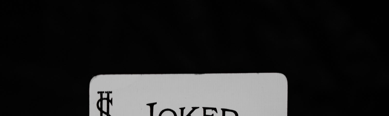 Black-and-white image on black background. Hand grasps partial deck of cards shown from the back, with joker card peeking out face-forward so that you can barely see his eyes — above his cap is the word “Joker.”