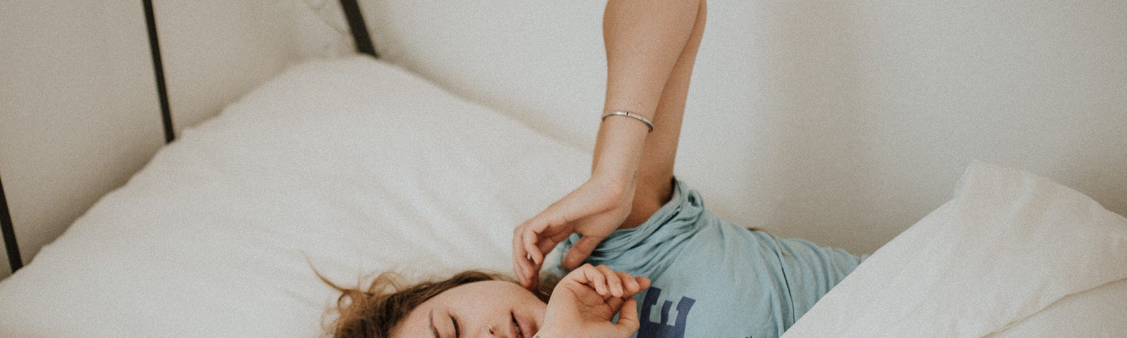 Photo of a woman in a blue t-shirt lying in a bed with white bedding. She has her eyes closed and her arms outstretched.