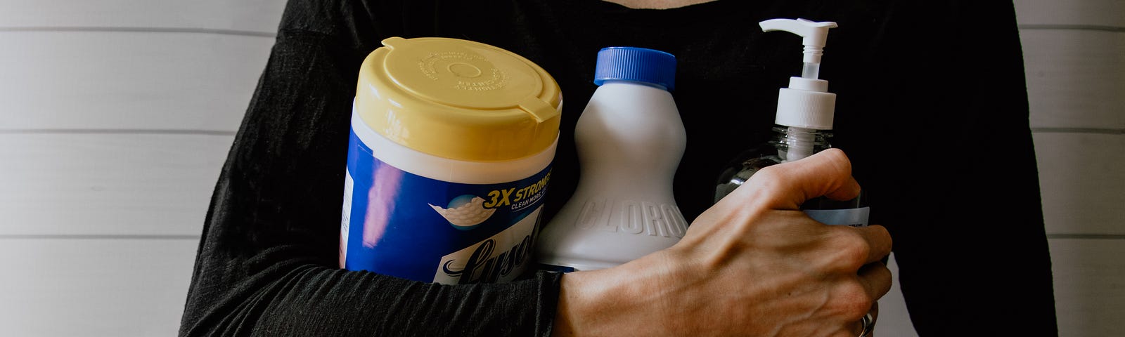 This is a photo that shows a woman from her waist to the base of her neck. She’s wearing a long sleeved black t-shirt, and she’s grasping 3 bottles of disinfecting cleaners.