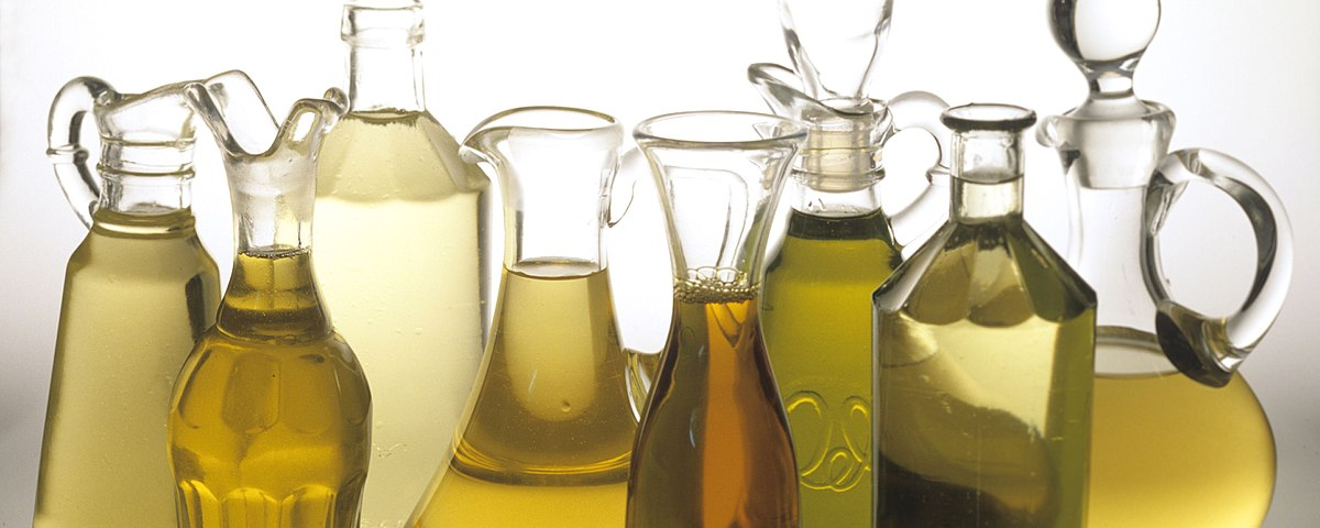 Five elegantly shaped glass pitchers with glass stoppers, two glass bottles, and a glass carafe. Some of the glass pitchers are curved and round while others have straight lines with curved handles. The bottles have unique scalloped edges that encircle the neck of the bottle with one bottle having more sharply defined edges than the other. They all contain oil with colors ranging from pale yellow, golden yellow, and amber.