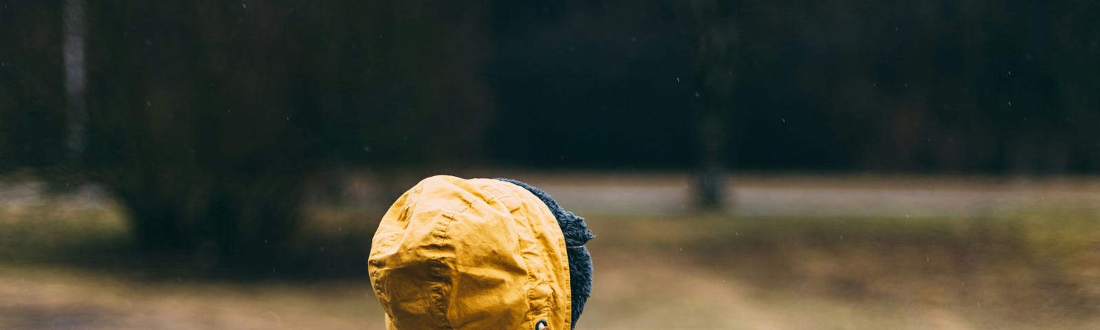 A toddler in a yellow coat with a blue backpack walking through the woods