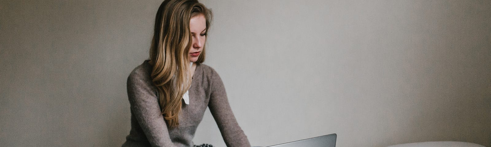 woman dressed for work, perched on single bed, typing on laptop