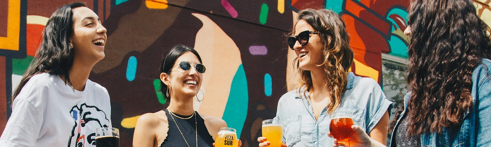 Four women in front of a mural sharing drinks, conversations, and smiles. It’s a thing!