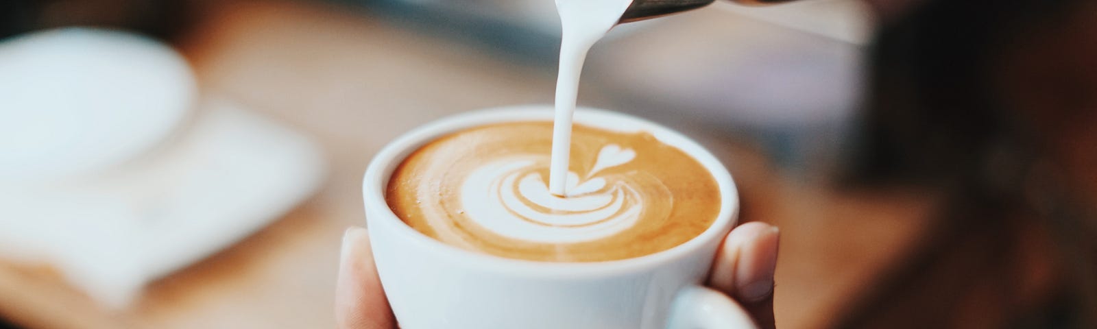 Photograph of milk being poured (out of a small steel container) into a small white cup. The pourer is creating a heart design in milk. Blurred background of a table with a cup and saucer.