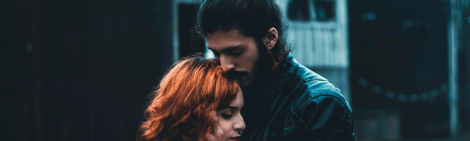 Man with black hair and a black leather jacket holding a woman with red hair.