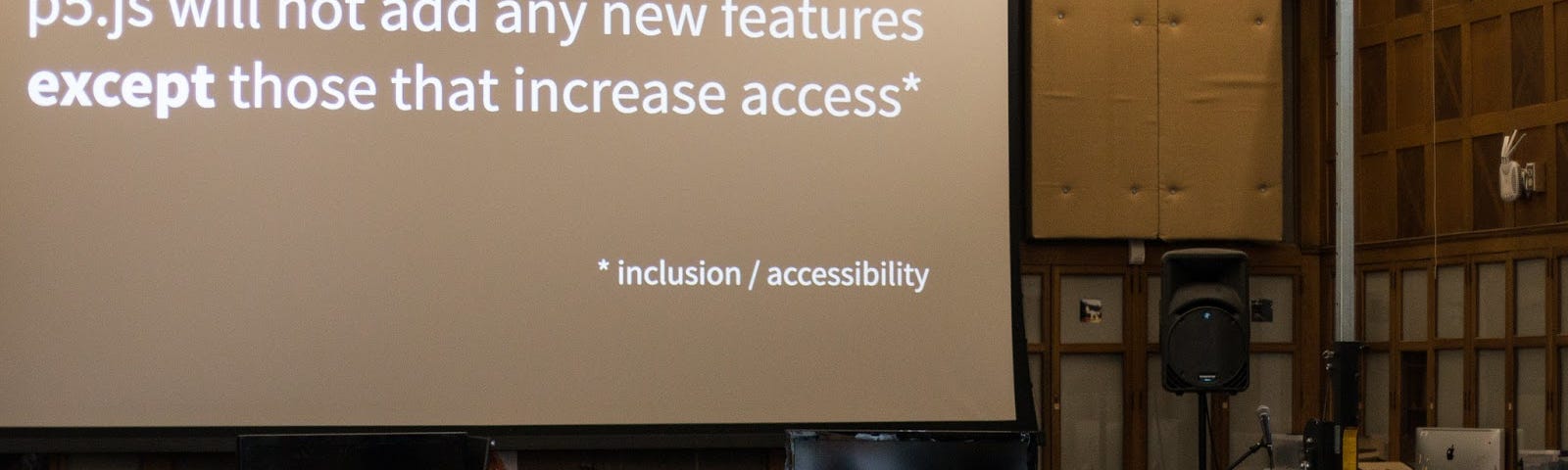 Group of people sit below a projected statement that says “p5.js will not add new features except those that increase access”