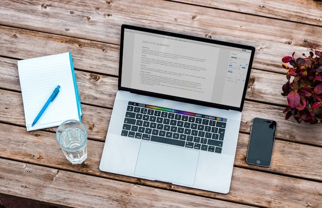 Top view of a laptop computer on a desk with a glass of water, a notepad with a pen, and a smartphone.